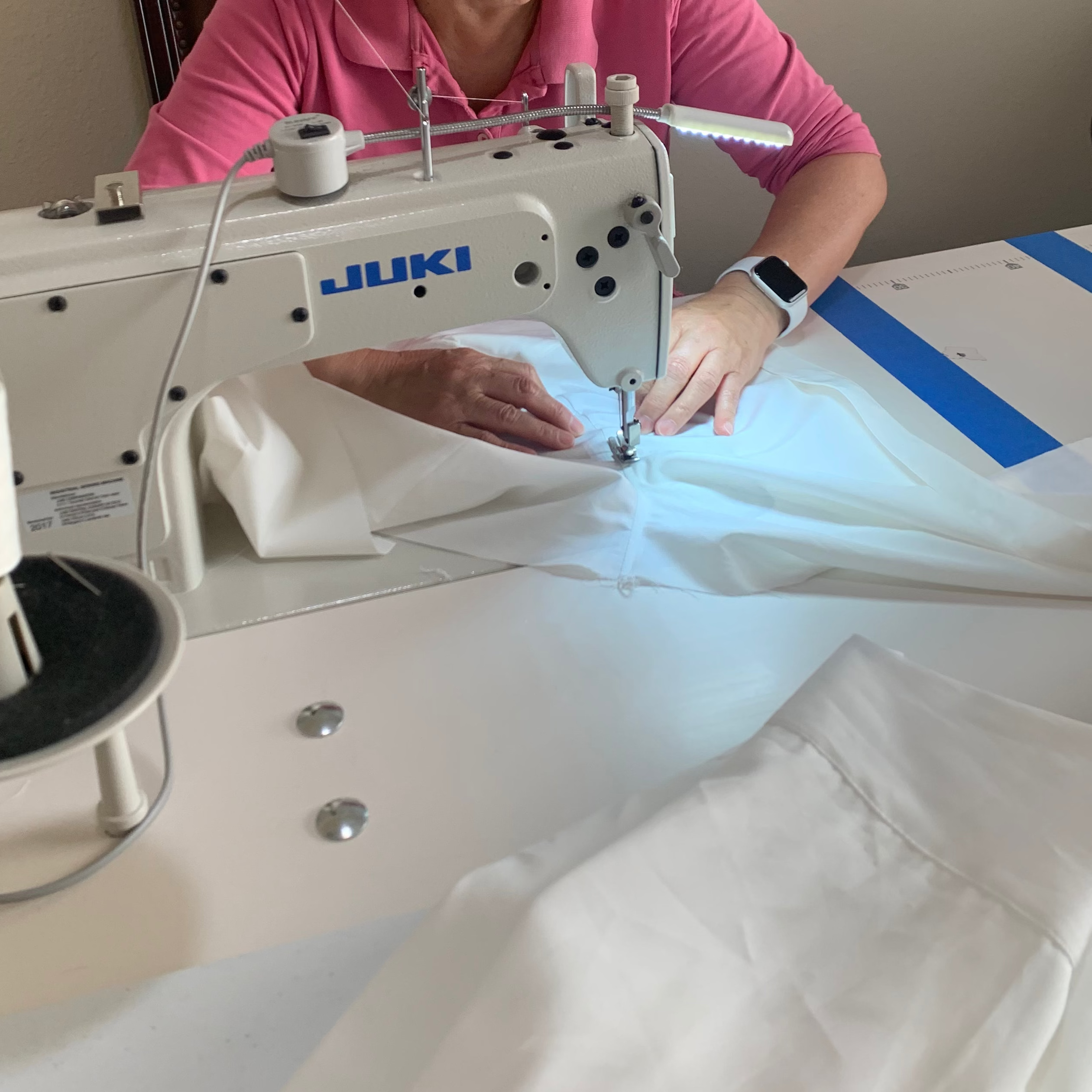 An older woman wearing glasses and a pink shirt operates a white JUKI sewing machine, stitching white fabric on a table.