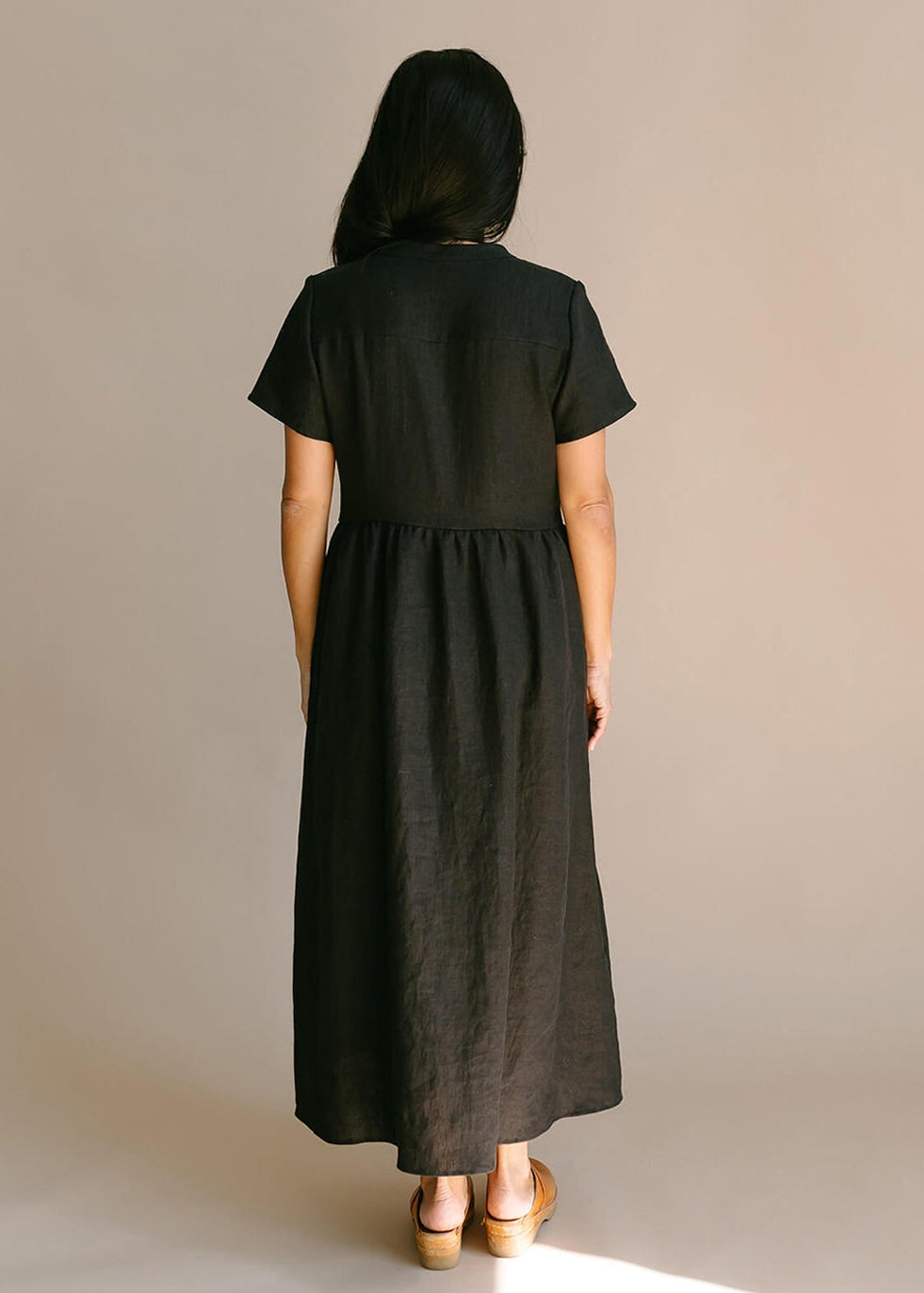 Back view of a a woman wearing a black, 100% linen, ankle length, v-neckline dress with fabric gathered at the waist, and two pockets.