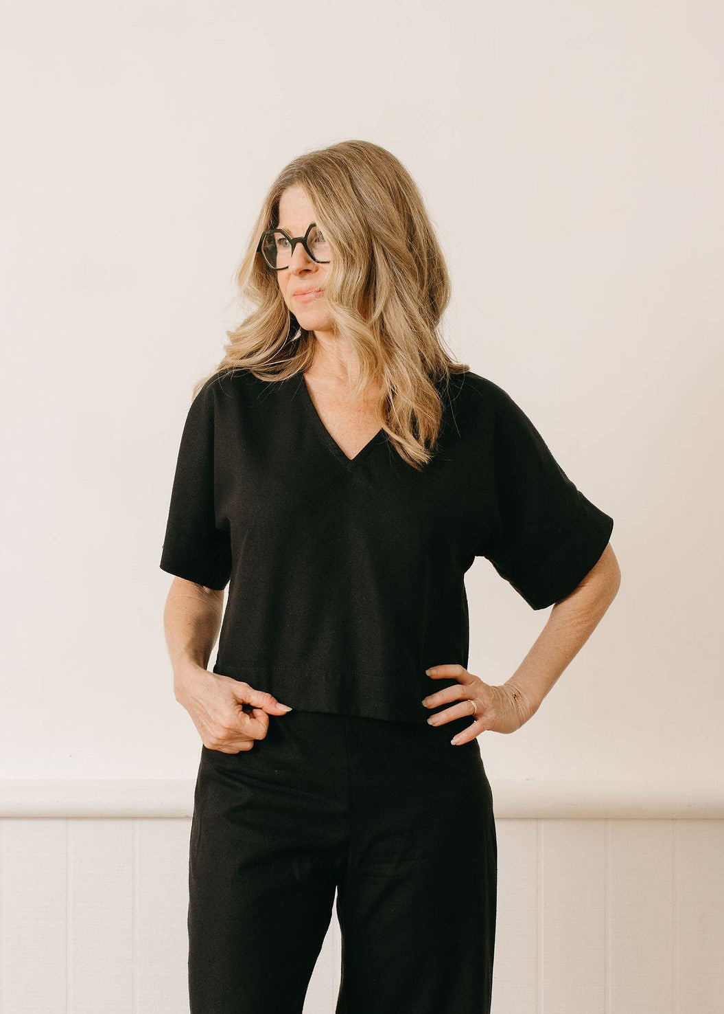 Woman wearing a black outfit with wide-leg pants against a white wall.