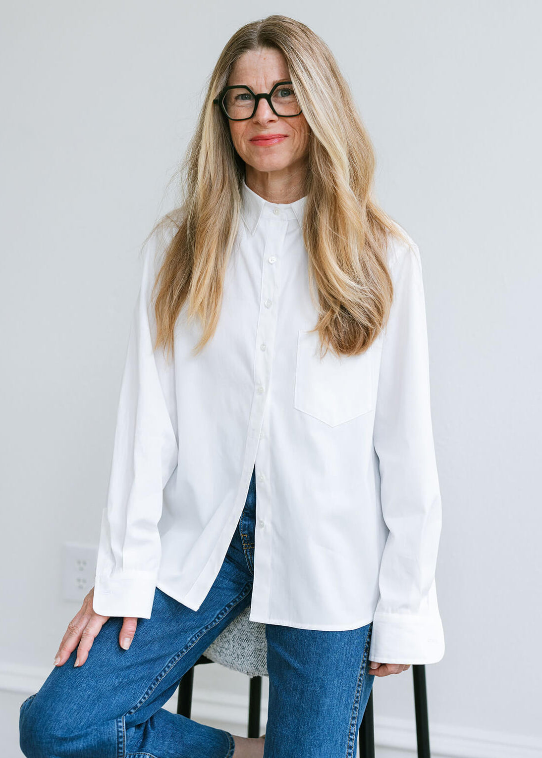 A woman with long blonde hair and black glasses wearing a white long-sleeve button-up shirt and blue jeans, sitting on a stool against a light gray wall.