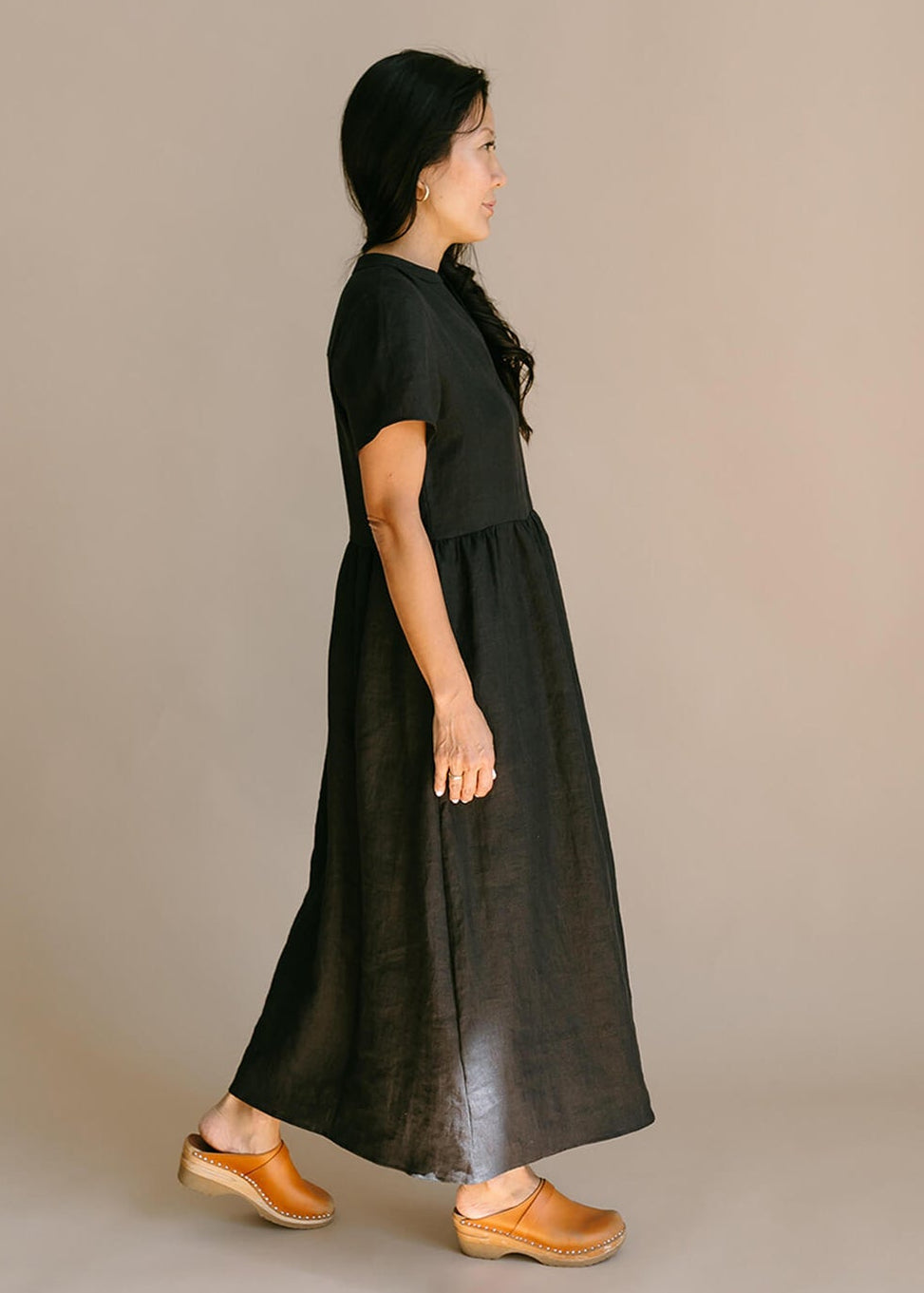 Side view of a woman wearing a black, 100% linen, ankle length, v-neckline dress with fabric gathered at the waist, and two pockets.