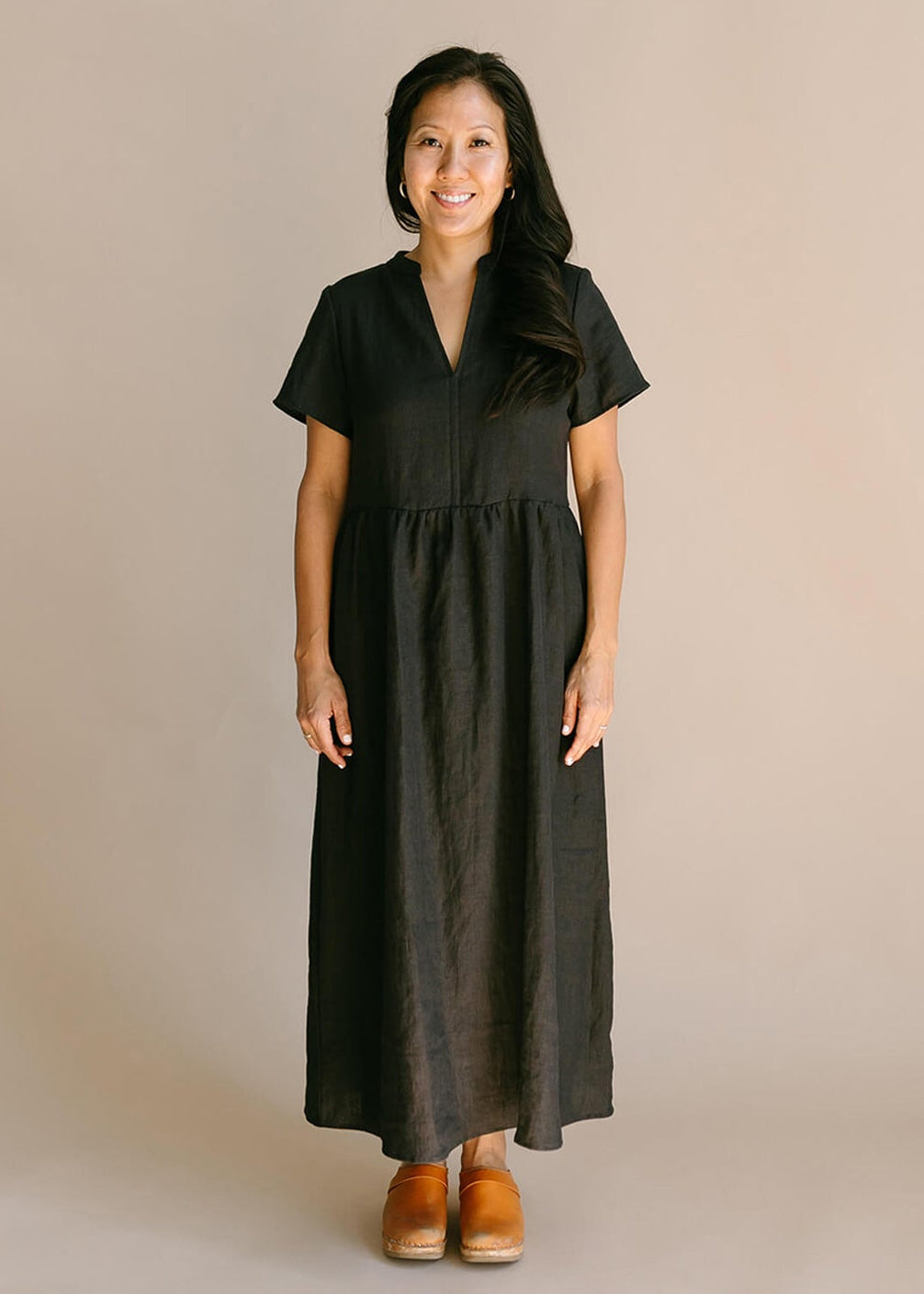 a woman wearing a black, 100% linen, ankle length, v-neckline dress with fabric gathered at the waist, and two pockets.