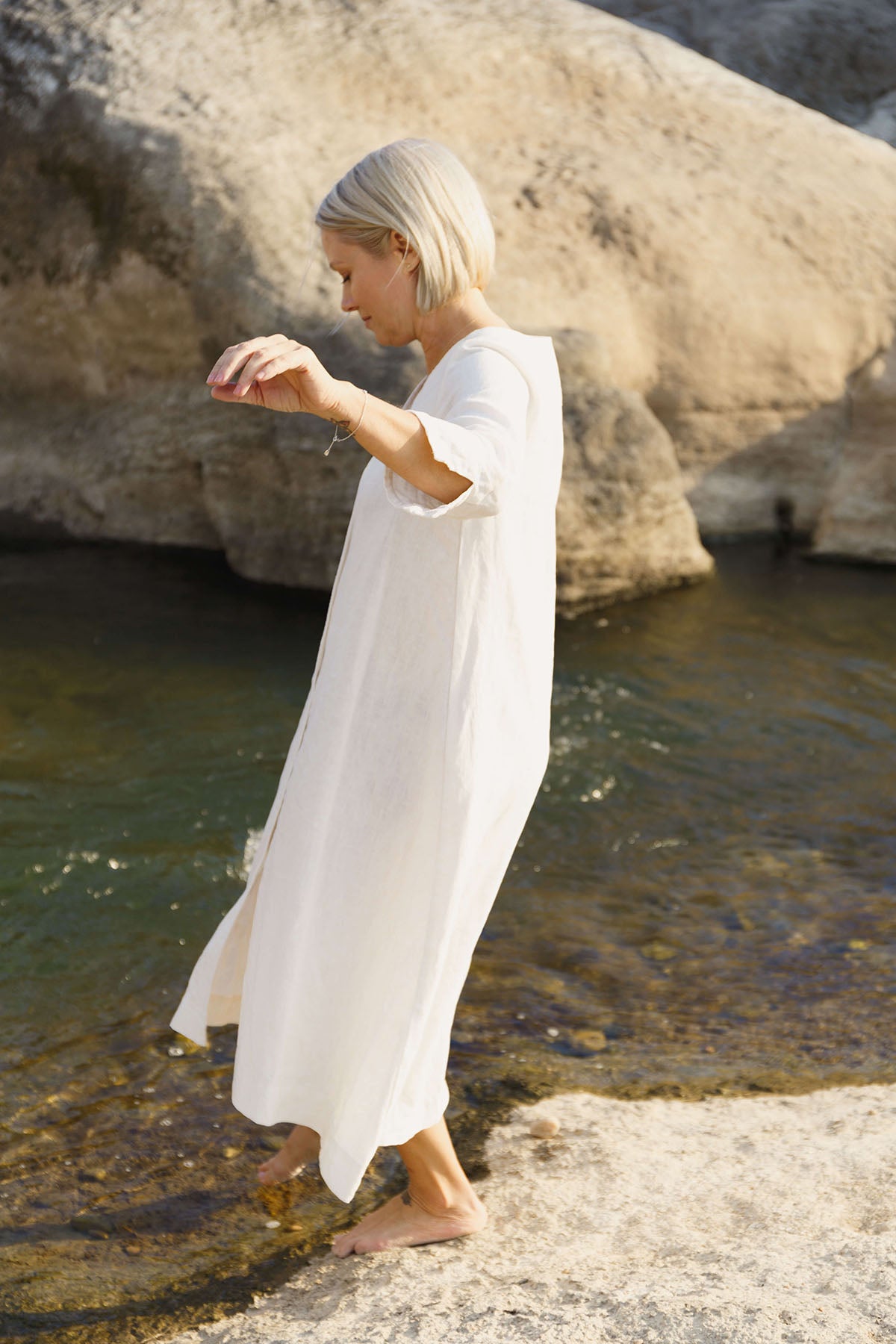 woman stepping into a creek wearing The Sienna Dress: everyday shirtdress with ¾ sleeves, a V-neck, an uneven hemline, made of 100% ramie.